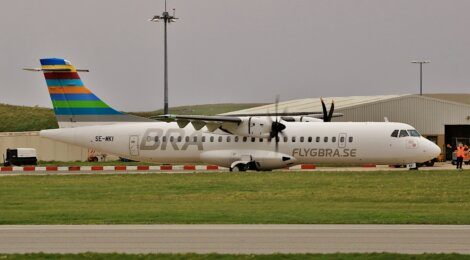 A FLYGBRA.SE commercial airplane with the registration SE-MKI is parked on an airport tarmac, with a terminal building and airport staff in the background.