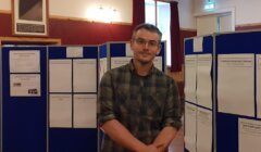 A man wearing glasses and a plaid shirt stands with crossed arms in front of blue display boards covered with papers in a room with wooden walls and red curtains.