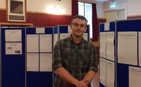 A man wearing glasses and a plaid shirt stands with crossed arms in front of blue display boards covered with papers in a room with wooden walls and red curtains.