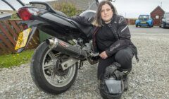 A person in motorcycle gear crouches beside a black motorcycle, holding a helmet.