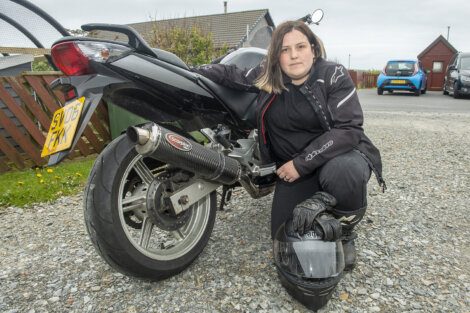 A person in motorcycle gear crouches beside a black motorcycle, holding a helmet.