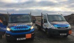 Two ZetGas branded vehicles parked on a rugged outdoor location under a cloudy sky. Both vehicles display logos featuring blue flames, with one being a flatbed truck and the other a van.