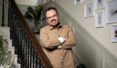 A man with glasses and a beard, dressed in a beige shirt, stands on a staircase with his arms crossed, smiling. Framed pictures hang on the wall in the background.