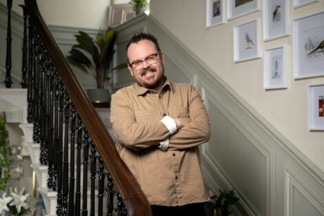 A man with glasses and a beard, dressed in a beige shirt, stands on a staircase with his arms crossed, smiling. Framed pictures hang on the wall in the background.