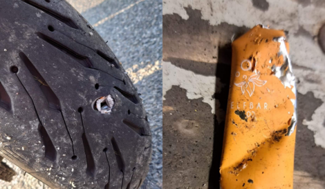 Close-up of a damaged motorcycle tire with a metal object lodged in it (left) and a burnt orange disposable vape labeled "Elf Bar" on the ground (right).