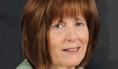 A middle-aged woman with shoulder-length brown hair and bangs, wearing a light green jacket and a necklace, poses against a dark background.