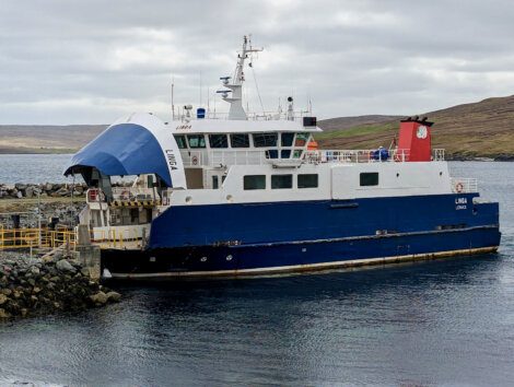 A blue and white ferry named "Linga" docks at a rocky pier in a calm coastal area under a cloudy sky.