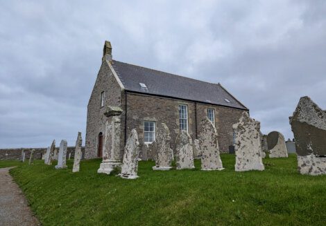 A small stone church with several old, weathered gravestones in front, set against a cloudy sky.