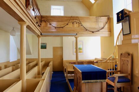 Interior of a small church featuring wooden pews, a blue-carpeted altar area, a second-floor balcony with netting, and simple decor including windows and framed pictures on the walls.