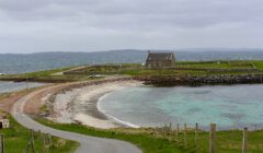 A narrow road leads to a stone house near a rocky shoreline with turquoise water and grassy landscape under a cloudy sky.