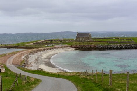 A narrow road leads to a stone house near a rocky shoreline with turquoise water and grassy landscape under a cloudy sky.