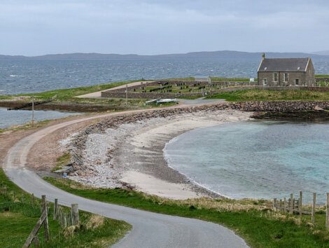 A coastal scene with a curved gravel road leading to a small stone house near the shore. A pebbled beach and clear blue waters are in the foreground, with distant hills visible across the sea.