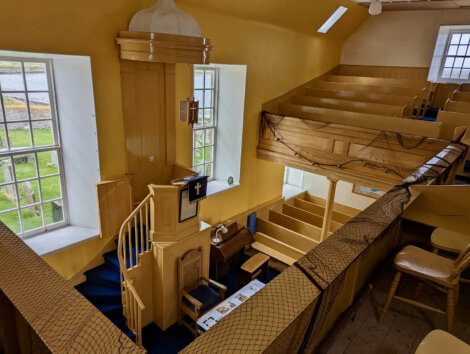 Interior of a yellow-painted church with a pulpit on the left, pews on the right, and a balcony. There are two large windows letting in natural light and a net strung along the balcony edge.