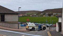 A police vehicle and officers are present at the entrance of a residential area with houses and hills in the background. A "School Patrol" sign is visible on the sidewalk.