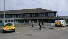 Two police vehicles and four officers are seen outside a building with a cordoned-off area. The officers appear to be securing the perimeter.