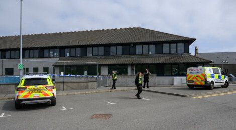 Two police vehicles and four officers are seen outside a building with a cordoned-off area. The officers appear to be securing the perimeter.