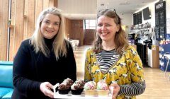 Two women smiling and holding a tray of assorted cupcakes in a bright, modern cafe with a wooden interior and menu board in the background.