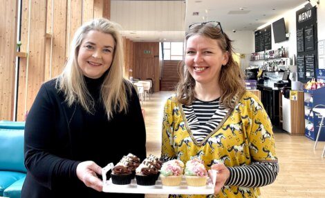 Two women smiling and holding a tray of assorted cupcakes in a bright, modern cafe with a wooden interior and menu board in the background.