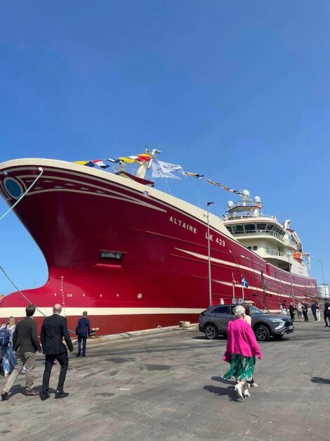 People walking towards a large red ship named Altaire LK 425 docked at a port on a sunny day.