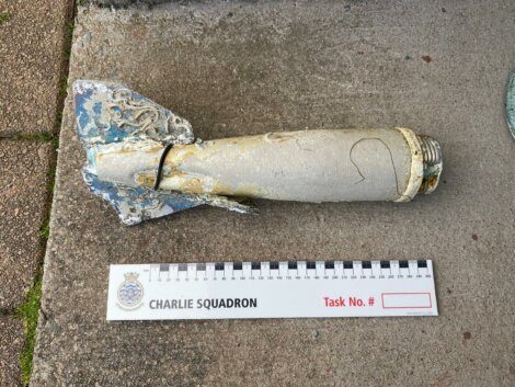 A damaged, corroded metal object resembling a missile or bomb is placed next to a measurement scale labeled "Charlie Squadron." The scale includes a task number section. The object is on a concrete surface.