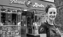 A woman stands smiling in front of a store named "Island Larder" which offers coffee, tea, hot chocolate, Shetland fudge, hampers, gifts, and other goods.