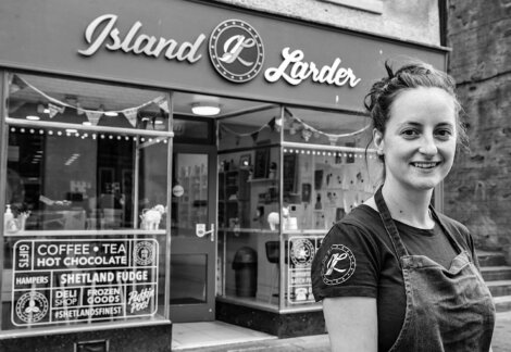 A woman stands smiling in front of a store named "Island Larder" which offers coffee, tea, hot chocolate, Shetland fudge, hampers, gifts, and other goods.