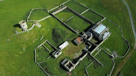 Aerial view of an ancient stone fortress with surrounding walls, partially intact structures, a circular tower, and grassy landscape.