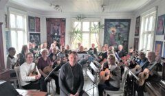 A group of people holding musical instruments, such as guitars and mandolins, are seated in a brightly lit room with artwork on the walls. Two individuals are seated at the front facing the camera.