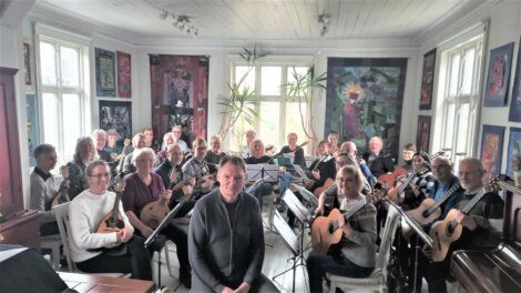 A group of people holding musical instruments, such as guitars and mandolins, are seated in a brightly lit room with artwork on the walls. Two individuals are seated at the front facing the camera.