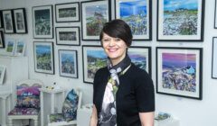 A woman, dressed in a black outfit with a floral scarf, stands smiling in an art gallery in front of various framed landscape paintings.