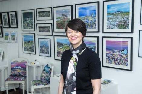 A woman, dressed in a black outfit with a floral scarf, stands smiling in an art gallery in front of various framed landscape paintings.