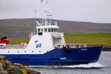 A blue and white ship named "BIGGA" is sailing near a rocky shore with hills in the background. There is a wind turbine visible on the hill.