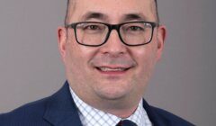 A man with glasses, wearing a blue suit, checkered shirt, and patterned tie, smiles against a gray background.