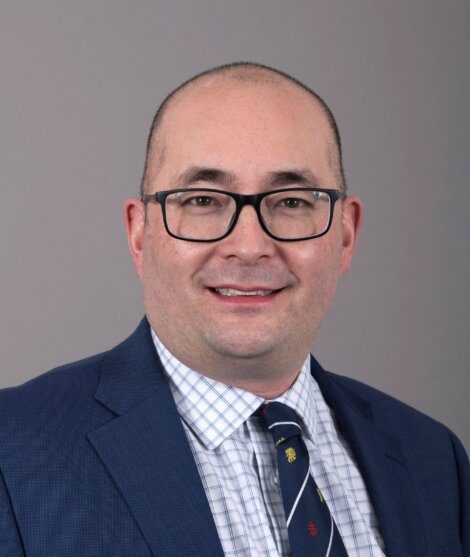 A man with glasses, wearing a blue suit, checkered shirt, and patterned tie, smiles against a gray background.