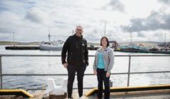 Two people stand in front of a harbor with a docked ship and buildings in the background on a cloudy day.