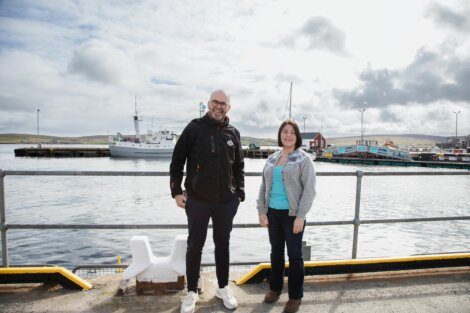 Two people stand in front of a harbor with a docked ship and buildings in the background on a cloudy day.