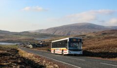 A bus marked "Toft 23" travels on a rural road surrounded by hills and scattered buildings under a clear sky.