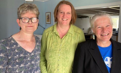 Three women standing indoors; from left to right, one in a floral dress, one in a green shirt, and one in a black jacket over a blue top. They are smiling and standing against a light-colored wall.