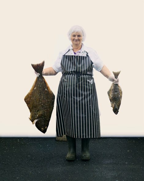 A person wearing a striped apron and hair net holds two large flat fish, one in each hand, standing on a black surface with a plain white background.