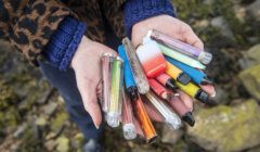 Person holding a collection of used disposable vapes in both hands, with a rocky background.