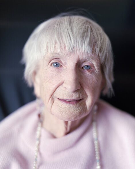 An elderly person with short white hair and blue eyes, wearing a pink top and a necklace, looking directly at the camera.