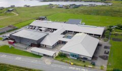 Aerial view of a school complex with multiple buildings surrounded by green fields, residential houses in the distance, and a body of water beyond. "Drop Off Only" is marked on the road near the entrance.