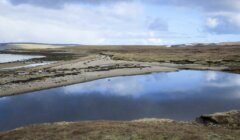 A calm lake reflecting the cloudy sky, surrounded by a barren, rocky landscape with sparse vegetation and distant hills.