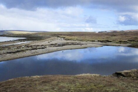 A calm lake reflecting the cloudy sky, surrounded by a barren, rocky landscape with sparse vegetation and distant hills.