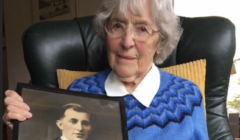 An elderly woman with white hair and glasses sits holding a framed black-and-white photograph of a man in graduation attire. She is wearing a blue and white sweater and is seated in a green armchair.