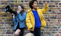 Two children stand against a brick wall; one, in a denim jacket, speaks into a megaphone, and the other, in a yellow jacket, holds a small protest sign and shouts. Another child is partially visible.