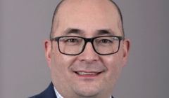 A man with short hair and glasses is wearing a blue suit, a white shirt with a checked pattern, and a blue tie. He is smiling in front of a plain grey background.