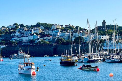 Boats float on the water near a marina with a town and hilly landscape in the background. Numerous sailboats are docked, and buildings are scattered on the hillside under a clear blue sky.