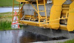 A yellow road construction machine spreads gravel onto freshly tarred road surface. A sign next to it indicates a speed limit of 20 mph and roadwork ahead.