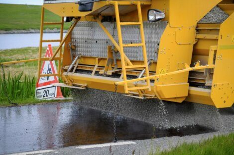 A yellow road construction machine spreads gravel onto freshly tarred road surface. A sign next to it indicates a speed limit of 20 mph and roadwork ahead.